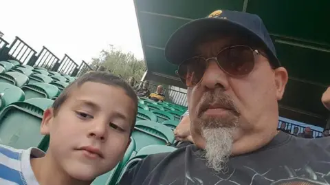 Child abuse training failures before Alfie Steele's Droitwich murder Family handout A man and a young boy sit in a sports ground that has rows of green chairs. The man is wearing a baseball cap and sunglasses, and he has a grey goatee beard. The boy has brown hair and he is wearing a blue-and-white striped T-shirt.