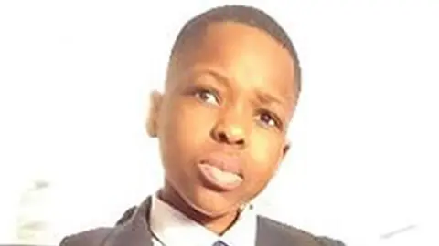 Met Police officer gives evidence Anjorin family Daniel Anjorin wearing school uniform of blazer, v-necked jumper, shirt and tie. He is looking toward the camera and is sitting on the back seat of a bus.