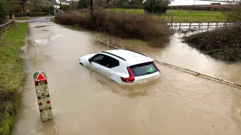 How to stay safe and what to do in a power cut Martin Giles/BBC A white car is stuck in flood water. The water is above the tyres and there is a measuring gauge at the side saying that the water is 3ft deep (0.9m)