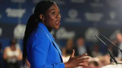 Kemi Badenoch calls for end to windfall tax on oil and gas companies PA Media A woman with long hair wearing a bright blue jacket making a speech standing in front of a lectern