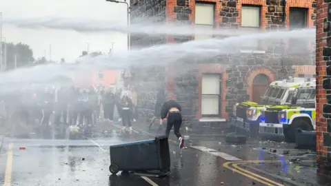 Ballymena sees second night of disorder with missiles thrown at police Pacemaker A group of people standing watching as someone in a tracksuit runs across a spray of water canon in front of a residential street and two police vehicles.