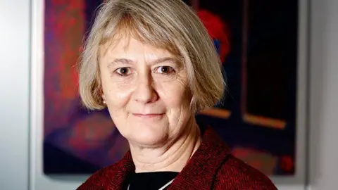 Foreign Office faces inquiry over its handling of Harry Dunn case Anne Owers with medium-length brown hair looking at the camera with a slight smile on her face. She is wearing a red jacket over a black top. She is standing in front of a red picture hanging on a wall.