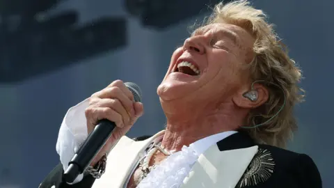 Old school charm from another era EPA Rod Stewart with a mic in his hand, throwing his head back on the Pyramid stage - he wears a white ruffled shirt and black jacket with gold embellishments