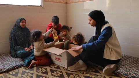 I feel like the life I had before the war was all made up Action For Humanity/Fadi Badwan Hanya sits on the floor, opening a cardboard box filled with aid supplies. Four children and another woman look on, two of the children retrieving items from the box.