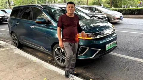 How China made electric vehicles mainstream Annabelle Liang Lu Yunfeng, a private hire driver, stands in front of his teal Dayun electric car that is parked along a road in Guangzhou in China. He is wearing a maroon t-shirt, striped grey pants and black shoes.