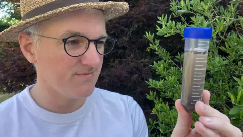 I found a bacteria-eating virus in my loo BBC James Gallagher wearing a white T-shirt and summer hat, looking closely at a vial containing pond-water