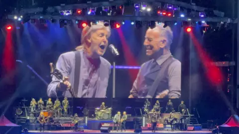 Bruce Springsteen and Paul McCartney in surprise Liverpool duet Sebastian Ayling A big screen and stage, showing Sir Paul McCartney signing into a microphone and holding a guitar. Bruce Springsteen is standing to his right holding a guitar and smiling, There is red and blue lighting on stage and there are other musicians.