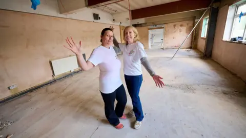 Leeds baby bank needs to raise £60,000 for new premises Leeds Baby Bank Two women wearing white t shirts and dark coloured trousers stand in a room which has bare plaster walls and floorboards, with their arms outstretched.
