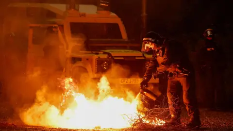 Twenty-nine arrests over recent violence Reuters Policeman in riot gear extinguishing a fire with a police landrover behind him.