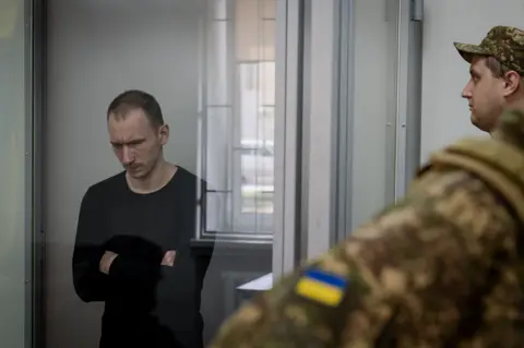 A Russian soldier takes the stand for an execution Dmitriy Kurashov, who has short dark hair and is wearing a black jumper, stands in the dock with his arms folded, looking down, surrounded by Ukrainian guards.