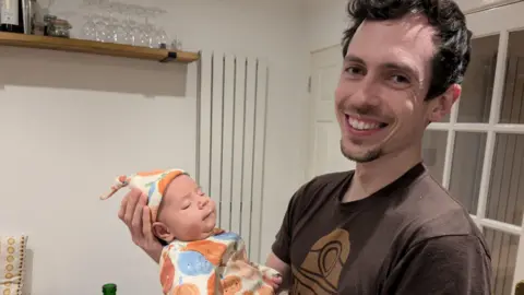 Paternity leave in UK is outdated and unequal, say MPs A man in a brown t-shirt smiles at the camera while holding his baby son