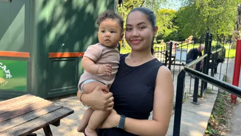 Why is Bristol's birth rate falling faster than other UK cities? Aaliyah standing in a Bristol park as she holds her baby in her arms. She is wearing a black dress and has dark brown hair tied back in a ponytail. She is smiling at the camera,