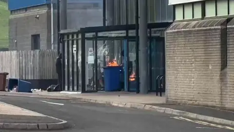 Police deploy more officers after 'mob violence' A blue wheelie bin being brought into a building, it is on fire.