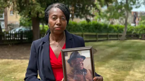 Covid inquiry begins examination of impact on social care sector Jean Adamson, wearing a red shirt and smart business suit, holding a photograph of her father Aldrick Adamson. Her hair is tightly plaited and tied back at the nape of her neck.