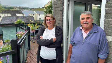 'The Heads of the Valleys road swallowed part of my garden' Julie and Daryl Wilkins stand outside Daryl's house, Julie with her arm crossed. He is leaning against the wall with the front door open beside him. Julie has chin-length blonde hair and is wearing glasses, a white t-shirt and a black jacket. Behind them decking can be seen with flower planters, with a row of terraced houses in the background.