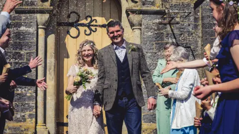 Glastonbury farmer 'makes more money from weddings than farming' River Lane Photography A bride and groom stand outside a church holding hands with guests looking on and throwing confetti. They are smiling and the bride is wearing a sequinned dress and holding a bouquet. The groom is wearing blue jeans, a brown tweed jacket, a navy buttoned-up waistcoat and a floral shirt.