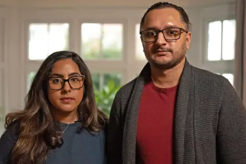 Leeds maternity services now 'inadequate' after inspectors act on parents' concerns MARTIN MCQUADE / BBC Amarjit and Mandip pictured standing next to each other. Amarjit has long brunette hair and black-rimmed glasses. She is wearing a blue jumper and a silver necklace. Mandeep has dark hair which is tied back, black-rimmed glasses and a short beard. He is wearing a red t-shirt and grey woollen cardigan. They are pictured in front of a white-framed window with green plants outside.