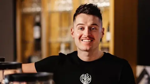 Uttoxeter coffee shop could shut over high rates, owner says Joe Ingleton A man in a black t-shirt. He is stood in a coffee shop next to two machines used to make coffee.