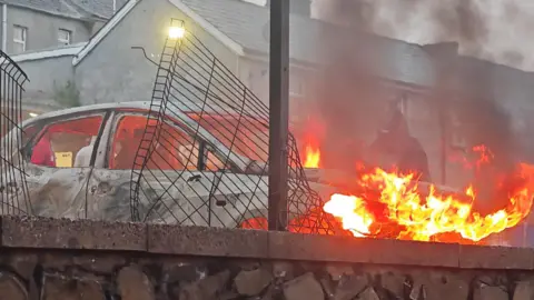 Ballymena sees second night of disorder with missiles thrown at police Pacemaker A white car has been set on fire outside a residential area. A hooded figure stands behind the orange flames.
