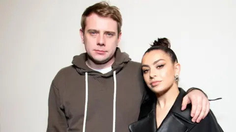 NI-born designer Jonathan Anderson becomes fashion brand's creative director Getty Images A man with light brown wearing a brown hoodie with white drawstrings and a white T-shirt underneath, looks into the camera. He has his left arm around a woman wearing her dark hair in a tight bun. She is wearing a dark top and large earrings.