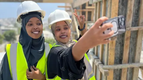 Somalia's construction boom in Mogadishu gives women high ambitions Anthony Irungu / BBC Saadia Ahmed Omar (right) takes a photo of herself and Fathi Mohamed Abdi (left) atop a building under construction in Mogadishu. They are both wearing hard hats over their headscarves and are in high vis yellow vests. Ms Omar makes the victory sign as she takes the photo.