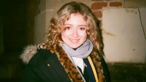 Our sister died because of our mum's cancer conspiracy theories, say brothers Gabriel & Sebastian Shemirani Paloma, pictured in what looks like a flash photo from a film camera, with her face brightly lit smiling in the centre of the frame, her hair in long blond curls, wearing a coat with a furry hood and with something in leopard print faux-fur around her shoulders. She is standing against a brick and stone wall at night