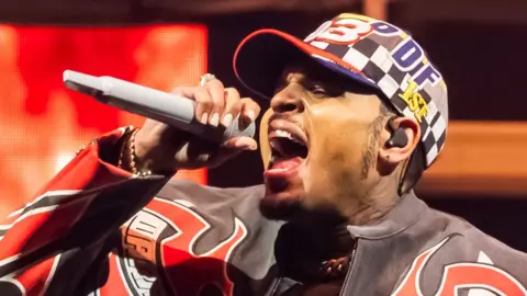 Chris Brown arrives in court to answer assault charges Getty Images Chris Brown in a red, white and black patterned leather jacket and patterned baseball cap, singing into a microphone on stage at the Tycoon Music Festival at Little Caesars Arena on April 19, 2025 in Detroit, Michigan.