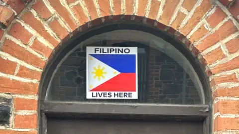 Police deploy more officers after 'mob violence' A sign reading "Filipino lives here" is stuck to a crescent window above a door, surrounded by the red brickwork of the front of a house. The sign also has the red, white and blue flag of the Philippines on it