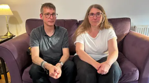'It was sold as a dream but became a nightmare' BBC Chris, a man with glasses and wearing a grey polo shirt, is sat on a purple sofa next to his wife Diana, who had long light brown hair and is wearing glasses and a white T-shirt. They are both looking at the camera