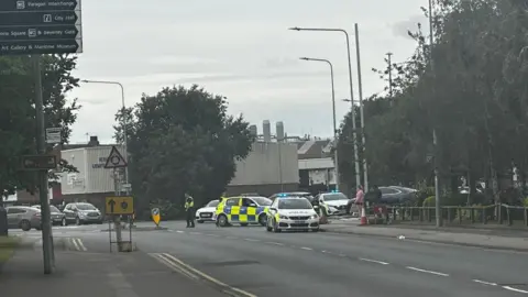 Ship on fire at Hull's Albert Dock as roads and shops are closed Amber Gash/BBC Three police cars block the entrance to a car park. A number of industrial buildings are in the background. A row of trees lines the road.