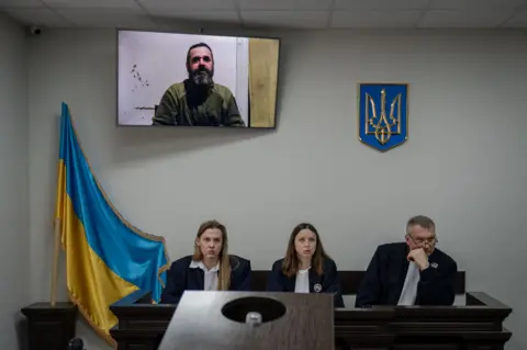 A Russian soldier takes the stand for an execution A Ukrainian soldier gives evidence on a television screen above a bench where three judges - two women and a man - are sitting