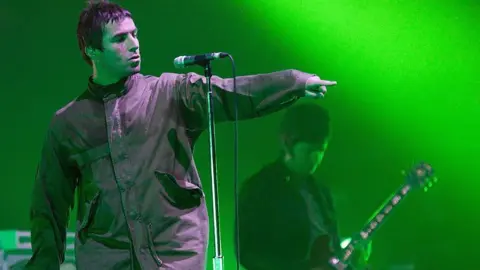 Ticket touts employing workers to bulk-buy for concerts Getty Images Oasis lead singer Liam Gallagher in a parka jacket pointing at the crowd in front of a microphone with his brother and bandmate Noel behind him, face down playing a guitar on stage in 2009 with green lights lighting the stage
