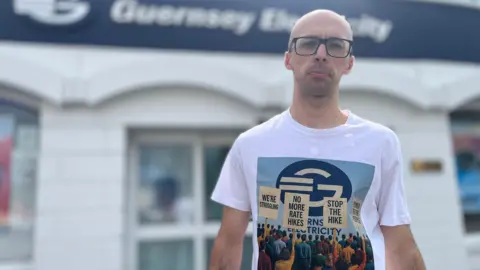 'We sit in the dark to save money on electricity in Guernsey' A bald man wearing glasses stands in front of Guernsey Electricity's head office. He is wearing a T-shirt with placards saying: "We're struggling! No more rate hikes, Stop the Hike and Power to the People."