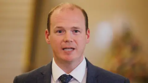 NI minister says scammers are targeting pensioners PA A balding man with blue eyes mid speech with a yellowy blurred background behind him. He is wearing a white shirt, navy tie with pink spots on it and a navy blazer.