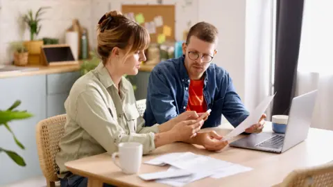 Council tax expected to rise by 5% a year Getty Images Man and woman sit at a kitchen table with a laptop, bills and a phone, with a noticeboard behind them.