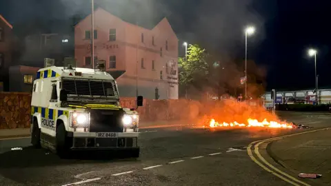 Twenty-nine arrests over recent violence PA Media A white police land rover with yellow and blue signage saying POLICE, parked in the middle of the road where a piece of material has been set a light across the road.