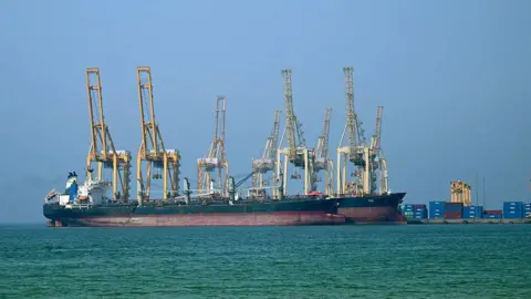 Turmoil and trade wars dominate 'summer Davos' Getty Images Two large ships with multiple cranes onboard are docked at a port in the background, with water in the foreground
