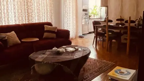 Trump's order to leave Tehran adds to fear as Iranians share 'last photo of home' Social media Living room with a sofa, coffee table and rug, with the curtains closed, and a dining table and kitchen in the background.