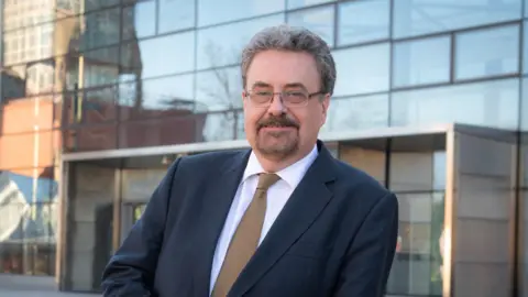 Dundee University bosses quit after damning report into financial collapse University of Dundee Prof Iain Gillespie in a blue suit and brown tie and wearing glasses, leans on a wall outside a university building