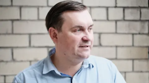 How Belarus dissidents in exile abroad are pursued and threatened Andrei Strizhak A man in a blue shirt and dark hair stands in front of a white brick wall.