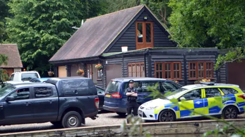 Police make renewed call over John 'Goldfinger' Palmer murder PA Media A uniformed police officer stands with his arms crossed beside a marked car, and surrounded by various other cars on the driveway area of John Palmer's home in South Weald. The picture has been taken a fair distance from the property.