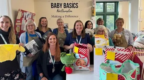 Wollaston charity supports one in 16 babies across the county Annabel Amos/BBC A group of women of varying ages, smiling at the camera. Some are holding clothes. toys and other items. Some are holding bags full of items. The words BABY BASICS appear in blue lettering on a wall behind them.
