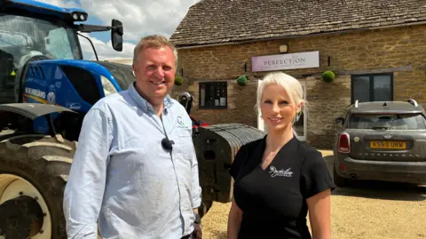 Glastonbury farmer 'makes more money from weddings than farming' A man in a pale blue shirt with blonde hair stands next to a woman with blonde hair in a black beauty tunic. Behind them is a tractor and a car parked outside a farm building with a pink sign on it.