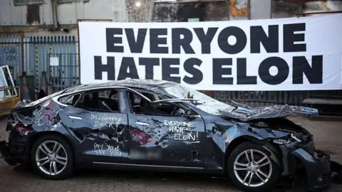 Will Musk's explosive row with Trump help or harm his businesses? Getty Images A banner reading "everyone hates Elon" behind a smashed up Tesla