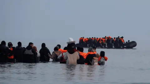 Smugglers deploy 'taxi boats' to collect migrants off the French coast Lea Guedj/BBC A group of people stand in waist-deep water with an overloaded inflatable boat in front of them