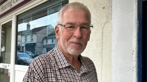 NHS and education named as local priorities A man, wearing a flannel shirt and glasses. He is looking at the camera with a slight smile. He is stood in front of a shop. There is a cream wall and windows and a white door. The logo on the shop says "the New Hearing Centre"