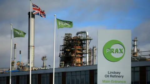 Calls for inquiry as hundreds of jobs at risk at Lindsey Oil Refinery Getty Images A metal structure in the background with a board on the left reading: 'PRAX. Lindsey Oil Refinery. Main Entrance'. There is a union jack flag on the left between two green flags with PRAX written.