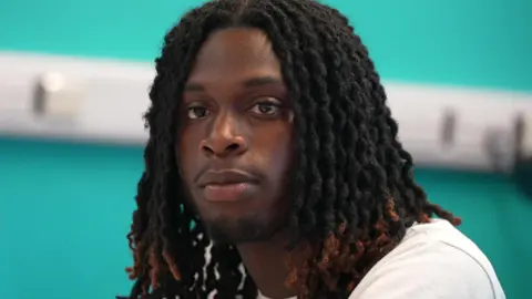 'Child Q case made me fear could this happen to me' Prince, 18, is wearing a white t-shirt, he is sitting at a table, with a bright blue wall behind him, taking part in the discussion