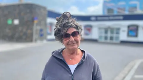 'We sit in the dark to save money on electricity in Guernsey' A woman with grey hair up in a bun, wearing sunglasses and a grey hoodie stands in front of Guernsey Electricity's showroom.