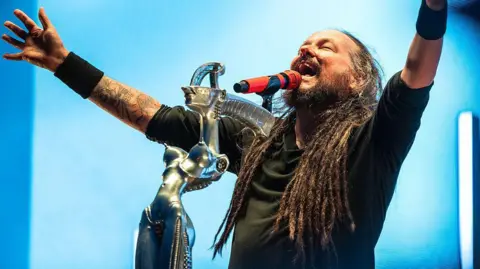 Y2K core is about nostalgia and escapism Getty Images Jonathan Davis, lead singer of the band Korn, holds his arms out in the air as he sings into a microphone on an elaborate stand in front of him. Behind him a bright blue screen lights up the scene.